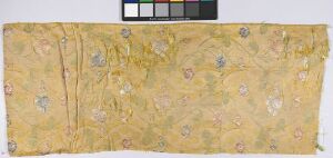  An aged, rectangular piece of textile with a muted yellow background, adorned with soft pastel-colored floral patterns in shades of blue, pink, and green, with visible creases and frayed edges, displayed against a neutral backdrop with a ruler for scale in the top right corner. Artistname and title are unknown.