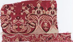  A detailed section of a rich burgundy textile featuring symmetrical, heart-shaped ornamental designs with intertwined elements in shades of red and accents of light beige.