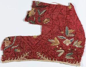  Fragment of an intricate red embroidered textile with gold, green, off-white, and brown floral patterns, showcasing sophisticated stitching and an irregular shape with frayed edges. Artist name and title of the piece are unknown.