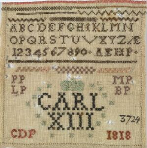  An antique, embroidered linen sampler with the Latin alphabet minus three letters at the top, two sets of numbers from 1 to 0 below, followed by a decorative pattern, and the name "CARL XIII" prominently featured in the center. Surrounding the name are