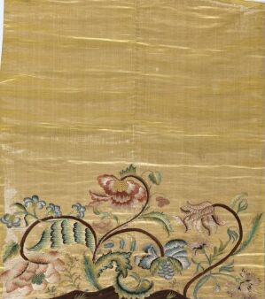  An image of a piece of vintage-looking embroidered fabric with a golden tan background and a pattern of soft-colored flowers and leaves stitched near the bottom edge, primarily in shades of green, blue, pink, and cream, on what may be a silk-like textile with vertical textured lines suggestive of aging or handling.