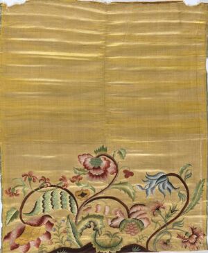  A photograph of a historical-looking golden-beige textile with vertical striations and intricate, botanical-inspired embroidery at the bottom. The design features stylized flowers in shades of pink, green, blue, and white, contrasting against the warm fabric.