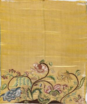  Embroidery on a faded yellowish-brown textile featuring a lower border of greens, blues, pinks, and purples in a floral and vine pattern, with evidence of aging and wear on the fabric.