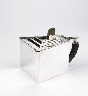  A modern silver teapot designed by Jacob Tostrup Prytz, featuring a geometric cube-shaped body, a flush flat lid with a ribbed handle, and a contrasting dark handle, set against a white background.