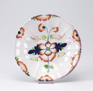  A decorative plate with scalloped edges featuring a symmetrical floral design with a central blue and white flower surrounded by green leaves, rust-colored flowers, and connected by a vine-like pattern on a white background.