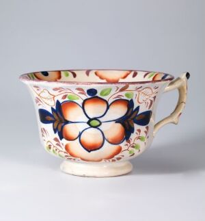  A colorful ceramic teacup with a handle, decorated with a large floral design in orange, peach, blue, and green hues, against a light gray background.