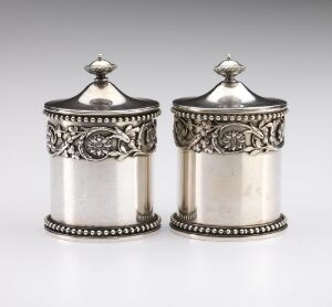  A pair of cylindrical silver containers with decorative bands and bead-like details, featuring domed lids with finials, set against a neutral gray background.