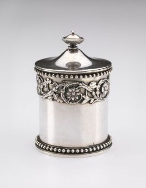  A small cylindrical silver container with an ornate top and embossed floral band, reflecting a soft gray metallic sheen on a plain background.