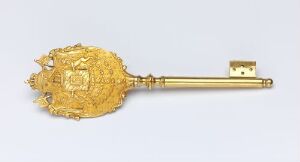  An ornate, gilded bronze key named "Kammerherrenøkkel" by an unidentified artist, featuring a sunburst pattern on the round handle with embossed and openwork decoration, leading to a smooth golden shank and a flat, notched end. The key has a shiny gold finish, indicative of ceremonial use or high-status function, set against a neutral background.