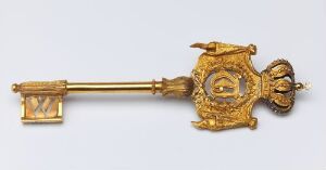  An ornate gilded bronze key known as "Kammerherrenøkkel" resting on a neutral background. The key features detailed patterns and a shield-like design in the center with a sculpted face on the bow, all finished in a shiny gold color indicative of its high status and ceremonial use.