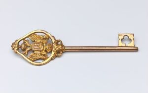  "Kammerherrenøkkel" by an unidentified artist, showing an ornate gilded bronze key with an intricately designed bow featuring a coat of arms and openwork decoration, set against a light-grey background.