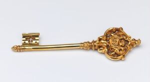  An ornate gilded bronze key known as 'Kammerherrenøkkel' by an unidentified artist, showcasing a complex, circular floral-like motif in the handle with partially openwork design, leading to a long, smooth shaft and a modestly designed bit against a light grey background.