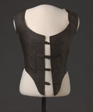  An upper torso mannequin with a dark-colored, possibly black, historical corset or bodice featuring structured ribs and decorative horizontal strips, set against a dark grey background.