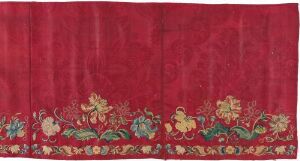  A fragment of an aged textile with a dark crimson background, showing wear and discoloration, adorned with a detailed and symmetrical border of stylized green, gold, and red floral and mythical creature motifs along the bottom edge.