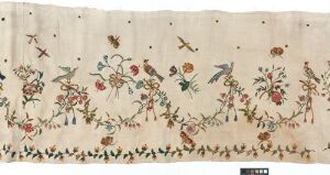  A detailed image of a beige or cream-colored embroidered textile featuring a repeating pattern of green, vine-like stems with colorful flowers and perched birds, rendered in red, pink, blue, orange, and brown, showcasing traditional embroidery craftsmanship.