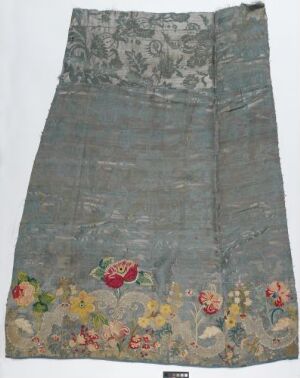  An antique-looking rectangular blue-gray fabric garment panel with faded gray upper design and vibrant, colorful floral embroidery towards the bottom.