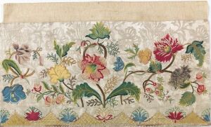  A vintage textile featuring an ornate pattern of colorful, detailed flowers and green leaves on an off-white background with a creamy hue, showing signs of aging, possibly from an embroidery piece.