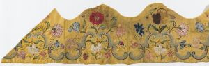  An elongated decorative textile panel with a scalloped upper edge featuring an intricate floral pattern on a goldenrod yellow background, with a large crimson flower surrounded by smaller blossoms in blues and pinks, and curling olive green stems with leaves.