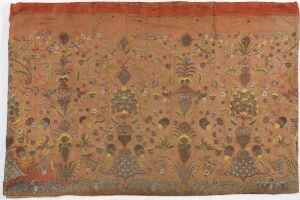  A rectangular piece of traditional fabric with a gradient background transitioning from burnt orange to mustard yellow to brown, decorated with repetitive patterns of stylized figures and motifs in dark brown and white, possibly indicating a form of traditional attire or ceremonial significance.