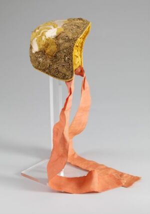  An ornate ceremonial hat with a textured gold body and patches of white, featuring a flowing coral pink ribbon, displayed on a transparent stand against a neutral grey background.