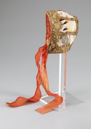  An artistic mask with gold and beige tones featuring hollow eye sockets and an open mouth, with flowing reddish-orange ribbon-like extensions, mounted on a clear acrylic stand. Artistname and title are unknown.