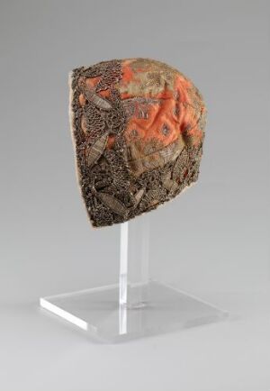  A textured cube with muted grays, browns, and vivid patches of orange-red on a display stand with a white base and transparent support against a light gray background. Artist name and title unknown.