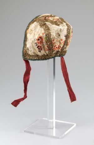  An intricately embroidered ivory cap with a floral pattern in red, pink, blue, and yellow, with green foliage accents and long red ribbons, set against a grey background on a white stand.