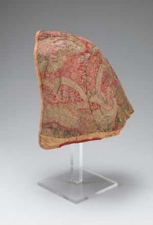  An ornate textile fragment with a red and beige damask pattern, mounted on a simple display stand against a neutral gray background.