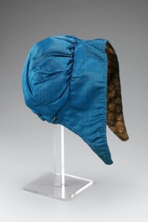  A brightly-colored blue satin bonnet with a fur-like brown and black animal print flap on one side, displayed on a white stand against a neutral background.