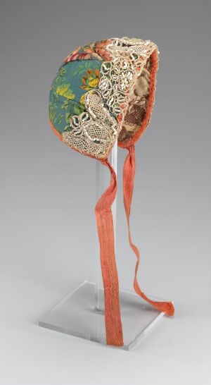  An art sculpture with a shiny, square metal base supporting a curved coral pink stem that holds a round ivory disc with a central lacy cutout, topped by a colorful mosaic resembling stained glass foliage.