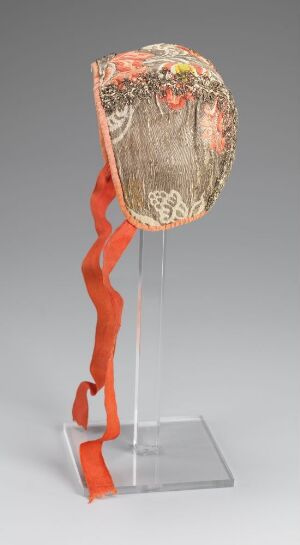  A sculpture featuring a circular element with ornate, faded designs in gold and orange on a cream background, mounted on top of a glossy, red, twisting ribbon-like support, presented on a transparent stand against a light grey background.