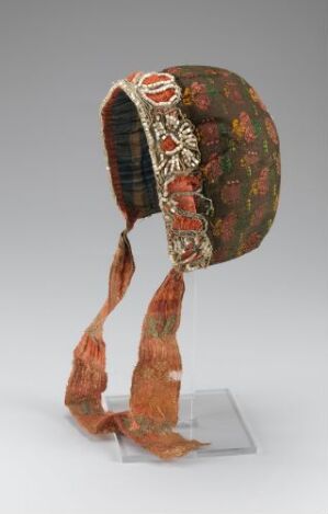  A traditional headpiece with a patterned fabric in shades of deep red, orange, green, and yellow, featuring a prominent ornamental badge with bright red and gold embroidery, and two decorative fabric straps hanging down.