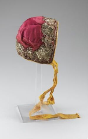  A historical or traditional cap with a deep pink top, elaborate gold embroidery, and long golden yellow trailing ribbons, displayed on a clear stand against a neutral background.