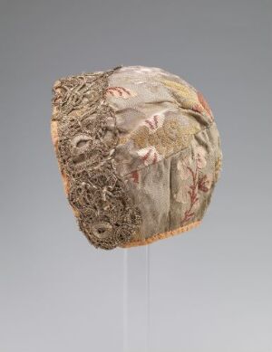  An intricately designed antique headgear with subtle hues of beige and pink, featuring layered fabric and lace, displayed against a neutral gray background with a clear supporting rod.