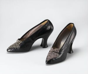  A pair of glossy black high-heel shoes with silver and gray floral lace detailing on the toes, displayed against a light gray background.