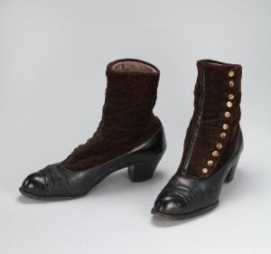  A pair of historical-style ankle-high boots with a plush chocolate brown body, black pointed toes, and heels, featuring a vertical row of decorative golden-brown buttons along the sides. They are displayed against a neutral gray background.