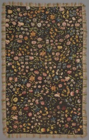  An intricately patterned textile with dense clusters of stylized flowers in warm tones of ochre, rust, gold, green, terracotta, pink, and yellow on a deep brown background, framed by light beige fringes on the edges.