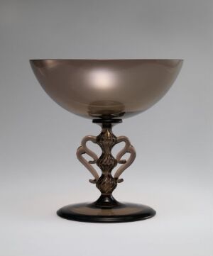  An elegant goblet with a broad, shiny bowl that transitions from silver to rose gold tones atop an ornate, dark bronze-colored stem with symmetrical, curvilinear shapes, set against a light gray background.