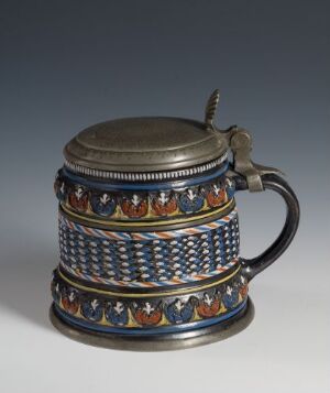  A traditional cylindrical beer stein with a hinged lid and handle, adorned with ornamental patterns featuring stylized red, white, and gold designs on a dark blue background, along with a smooth dark metal lid and handle.