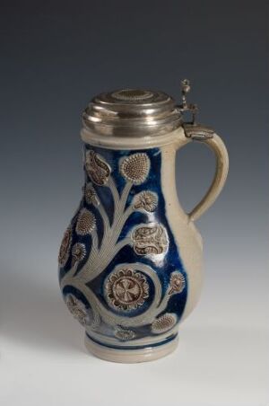  A stout ceramic jug with a metal lid, featuring a rich cobalt blue glaze and decorated with white swirling patterns and colorful heraldic medallions. The jug has an ergonomic handle and displays intricate craftsmanship. Artist and title are unknown.