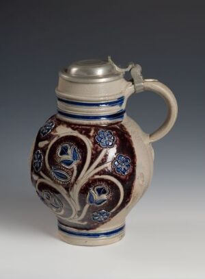  A ceramic jug with a white glaze, decorated with cobalt blue and dark brown patterns of foliage and rosettes, featuring a metal rim and lid, set against a soft gray background.