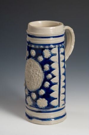  A cylindrical ceramic beer stein with a handle, featuring a cobalt blue and white design with embossed circular medallions surrounded by studded accents and spike-like decorations on a neutral gray background.