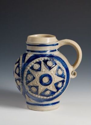  A ceramic jug with a creamy off-white base color and rich cobalt blue geometric and spiral patterns, featuring a central six-pointed star design. The jug has a sturdy off-white handle and a broad circular opening with a blue-trimmed lip, all set against a neutral gray background.