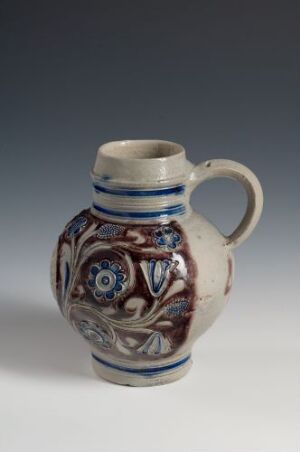  A small ceramic jug with a creamy white base color. It features bands and floral patterns in various shades of blue and brown, has a single handle, and is photographed against a neutral gray background.