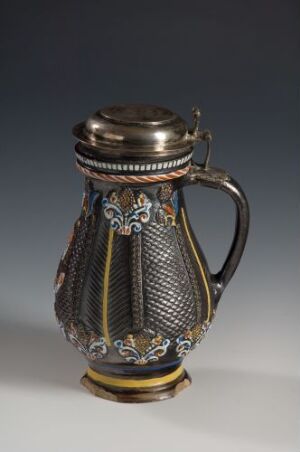  An intricately patterned metal tankard with blue enamel bands around the top and bottom, adorned with white and gold floral motifs, and a curved handle attached to its sides.