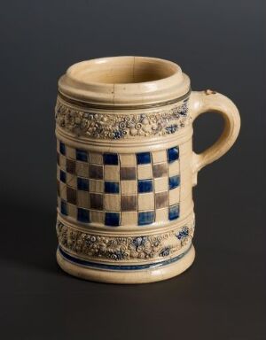  A traditional ceramic beer stein with checkered blue and cream patterns, adorned with metallic gold accents, displayed against a dark background. Artist and title unknown.