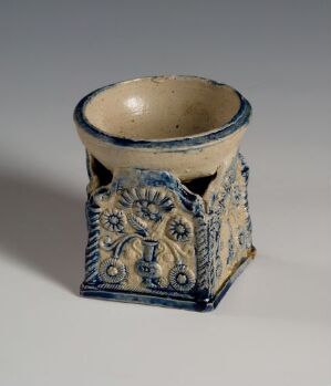 
 A small antique-looking ceramic vessel with a square base and a circular top, featuring cobalt blue floral embossing on a creamy white background, displayed against a light grey surface.