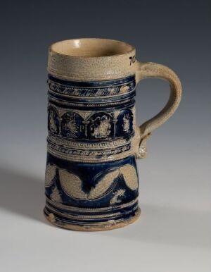  A historic-looking ceramic mug with a cream-colored base and deep cobalt blue decorative bands featuring scrollwork and scenes with figures and animals. The mug has a glossy finish on the decorative areas and a matte upper section and interior.