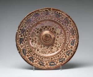  A ceramic plate with intricate brown, green, blue, and peach patterns displayed on a transparent stand against a neutral gray background. The plate features a central motif surrounded by concentric bands of decorative paisley and floral designs, culminating in a serrated edge.