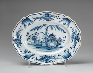  An antique oval-shaped ceramic plate with scalloped edges, showcasing a cobalt blue pastoral scene with a waterwheel and trees, set against a white background and framed by a decorative floral border. The plate has a rich historical ambience and is displayed against a light grey backdrop.